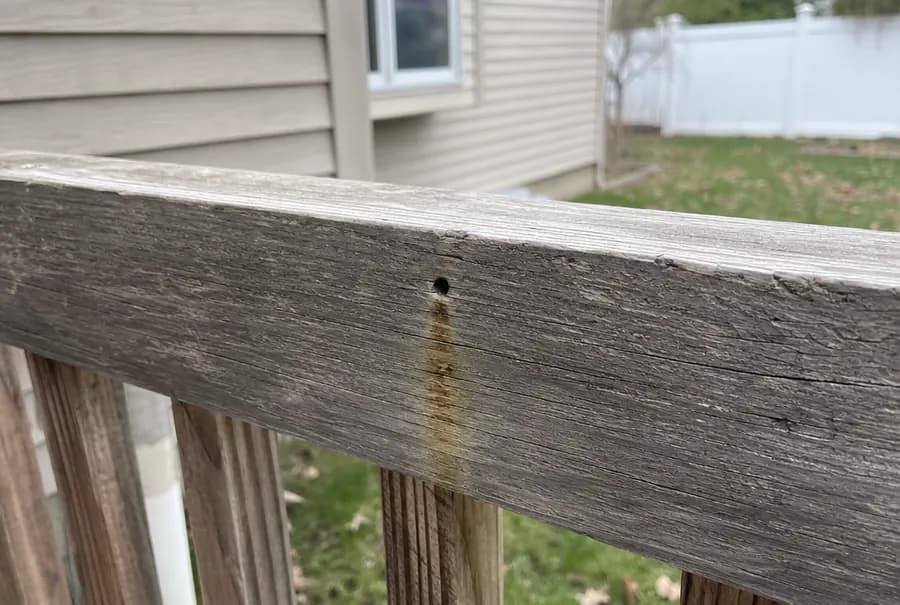 A yellowish brown stain streak running down a weathered deck rail below a small round hole