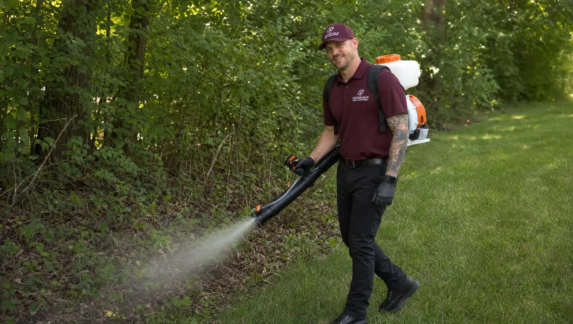 Honorable Pest Control technician in a burgundy uniform and black gloves misting the brush and leaf litter where a Michigan backyard meets the woods